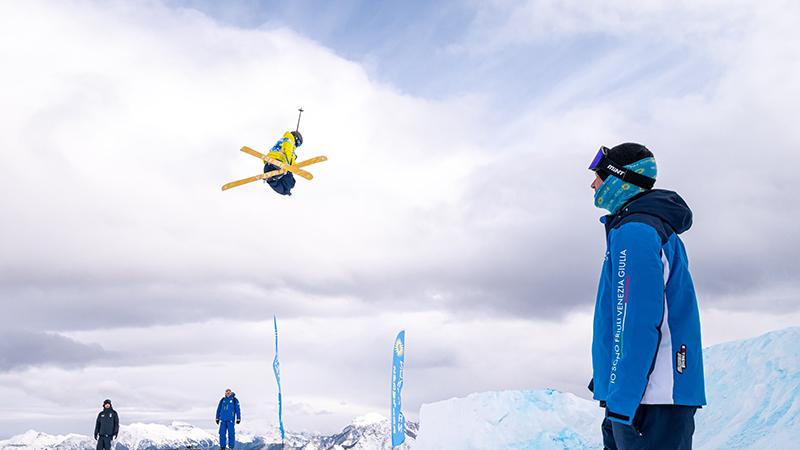 Ludvig Söderman hoppar högt med sina skidor under Europeiska Ungdoms-OS i Italien. I Förgrunden står en av tävlingens funktionärer och tittar på. 